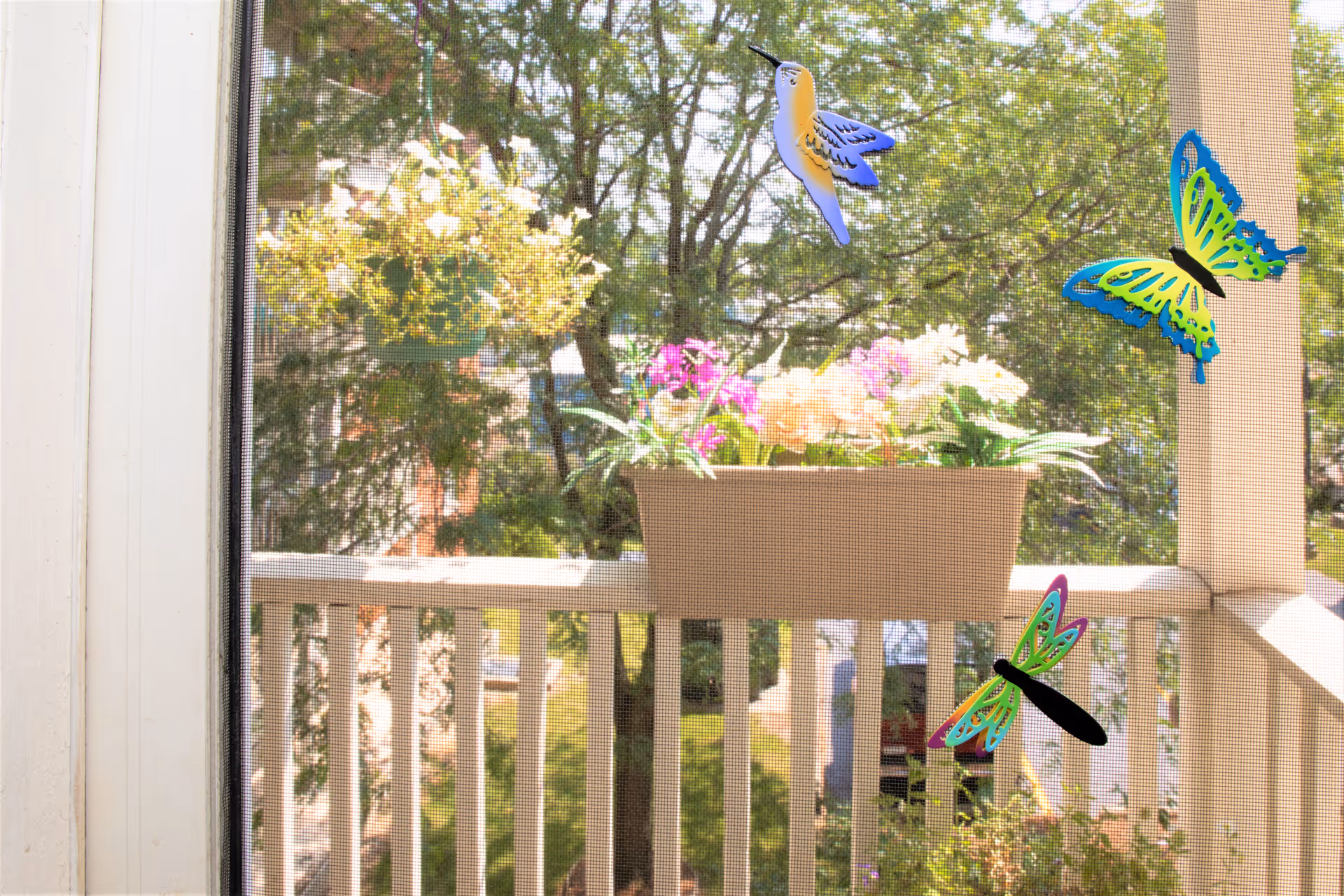 View through a screened window showing a balcony railing with a flower box containing colorful flowers. Decorative stickers of a hummingbird, butterfly, and dragonfly are attached to the window screen. Trees and greenery are visible in the background.