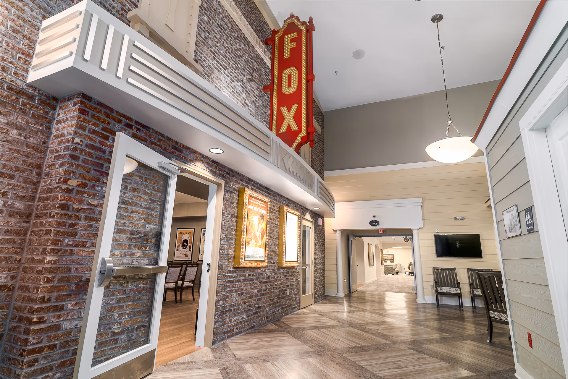 Interior hallway of The Memory Center featuring a brick wall with a large vertical red sign that reads 'FOX'. There are two illuminated movie poster frames on the wall and an open door leading to a room with chairs and framed pictures. The hallway has a high ceiling with a hanging light fixture, a television mounted on the right wall, and several chairs along the wall.