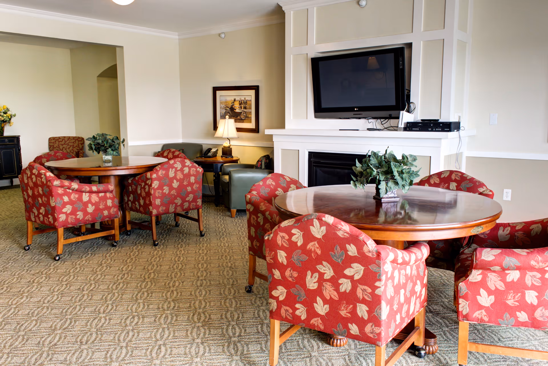 A cozy common area in a senior living facility featuring two round wooden tables surrounded by red upholstered chairs with a leaf pattern. In the background, there is a wall-mounted flat-screen TV above a white fireplace, a side table with a lamp, and a framed picture on the wall. The room has beige walls and carpeted flooring with a geometric pattern.