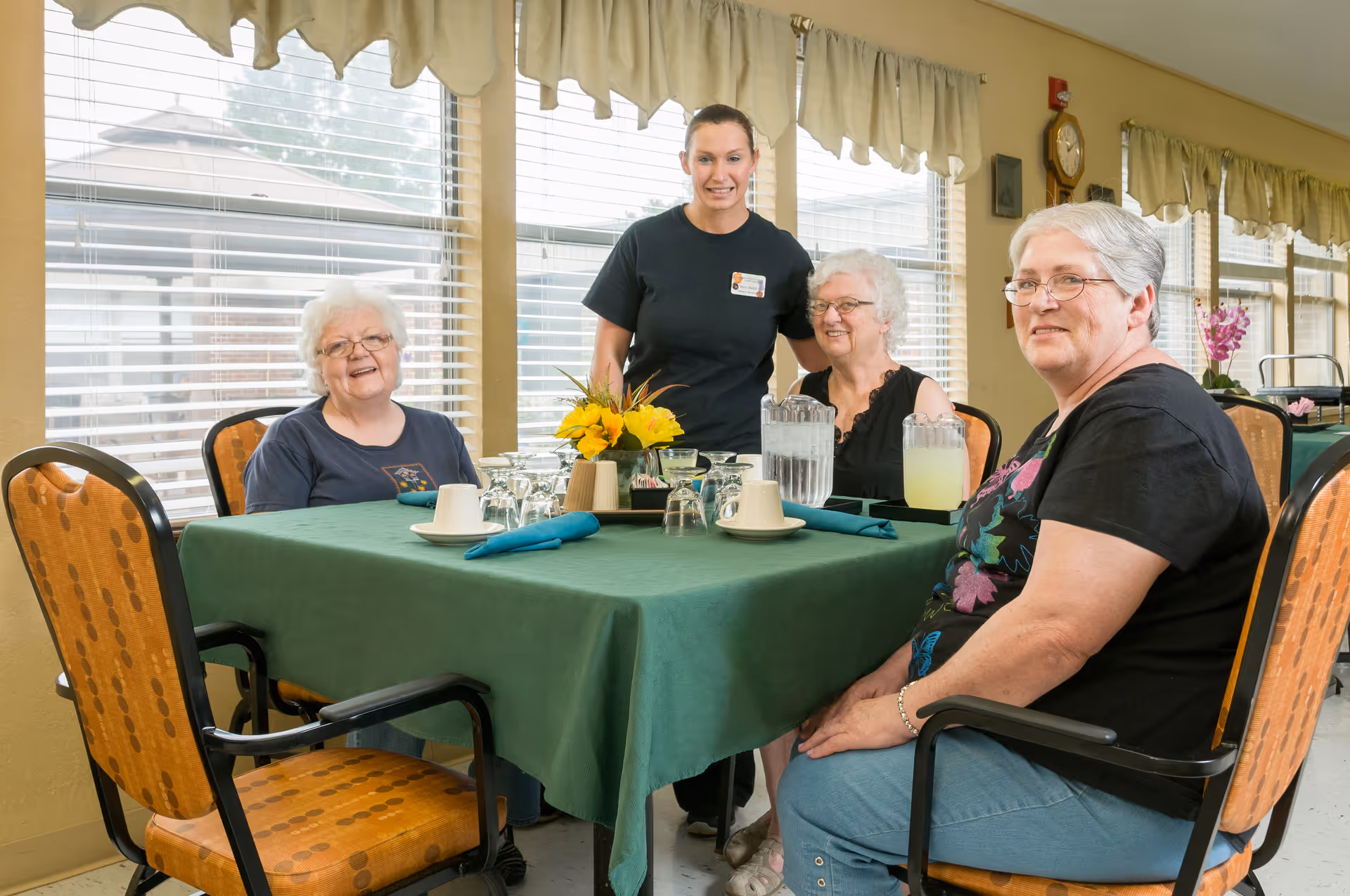 Three elderly women seated around a dining table with a green tablecloth, set with cups, glasses, and pitchers of water and lemonade. A caregiver stands behind them smiling. The room has large windows with blinds and beige curtains, and the setting appears to be a communal dining area in a senior living facility.