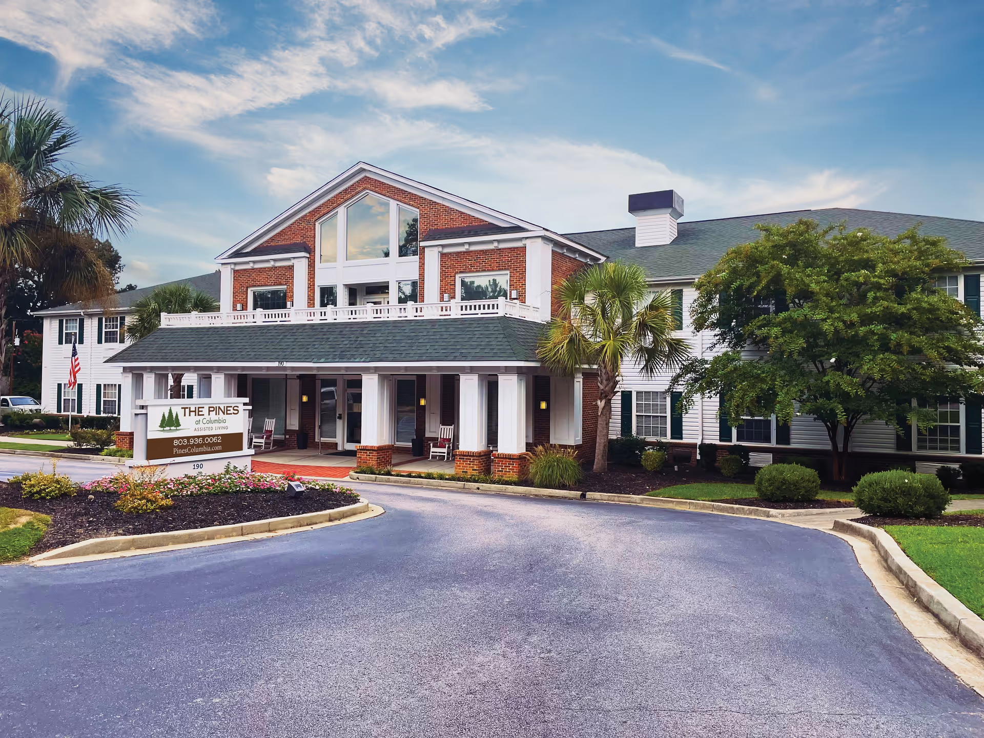 Front exterior view of The Pines at Columbia Assisted Living Community building with a driveway, landscaped garden, palm trees, and a sign displaying the facility's name, phone number, and website.