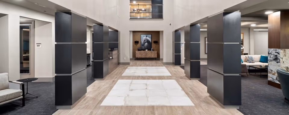 Spacious and modern interior common area with tall black pillars, light wood and tile flooring, and seating areas with sofas and chairs on either side. A large framed picture of a white horse is displayed on the far wall above a console table.