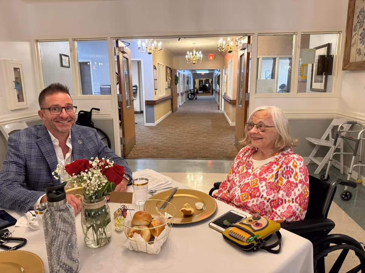 An elderly woman in a wheelchair and a man smiling across a table with flowers, rolls, and plates in a senior living facility dining area.