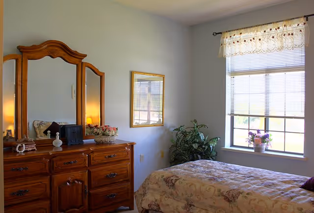 A cozy bedroom with a wooden dresser featuring a three-panel mirror, decorative items, and a floral arrangement. A bed with a floral bedspread is positioned near a window with blinds and a lace valance. A potted plant is placed in the corner near the window.