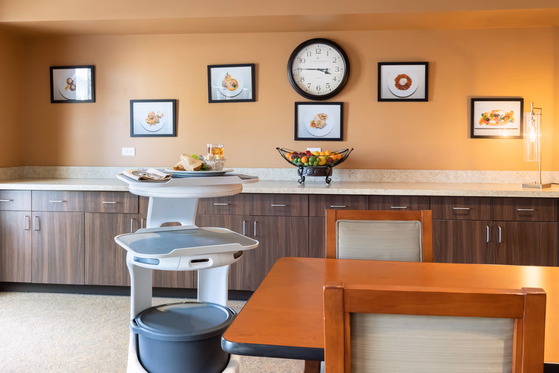 Interior view of a dining area in a senior living facility with a wooden table and chairs in the foreground. A serving cart with food and drink is positioned near the table. The back wall features wooden cabinets with a light countertop, a clock showing 3:45, and six framed pictures of plated food dishes. A bowl of fruit is placed on the countertop.
