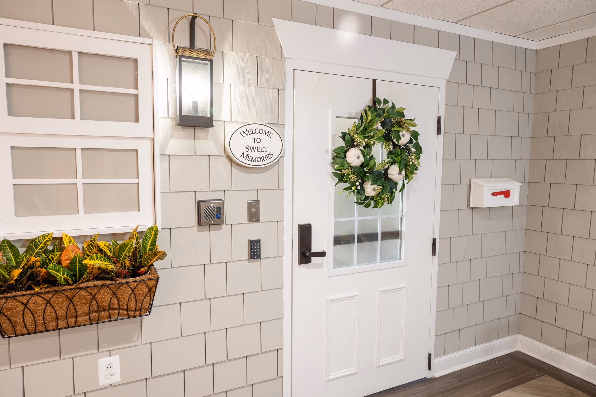 Interior entryway with a white door decorated with a green wreath, shingle-style walls, wall lantern, planter of greenery and a 'Welcome to Sweet Memories' sign.