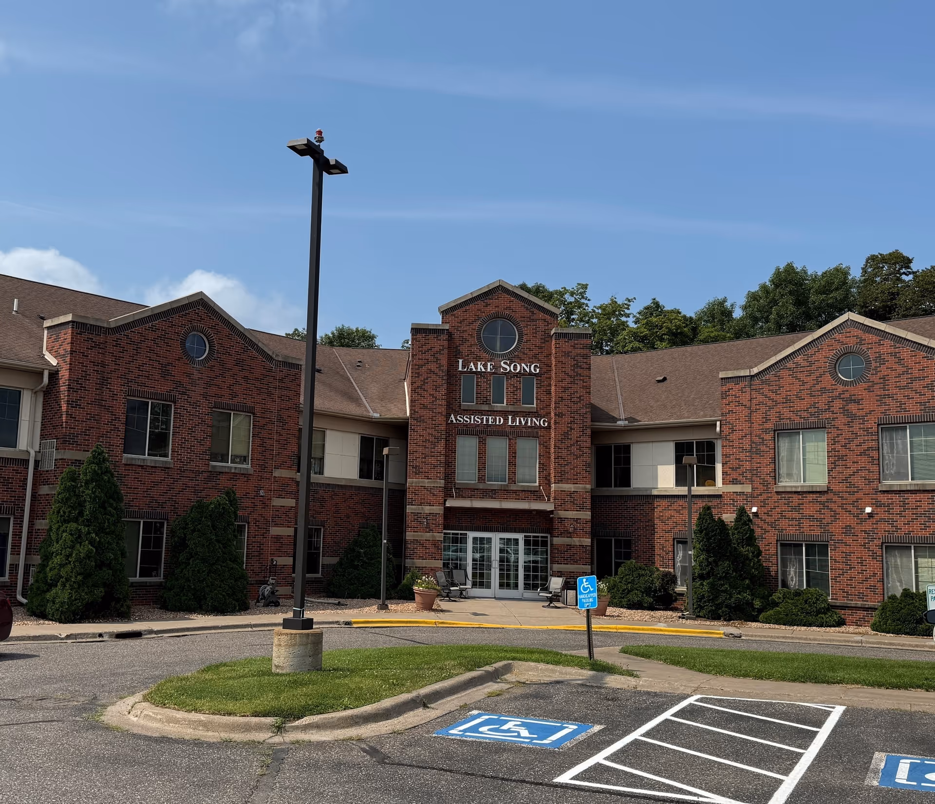 Exterior front view of Lake Song Assisted Living facility, a two-story brick building with multiple windows, a central entrance with glass doors, and a parking area with handicapped parking spaces in front.
