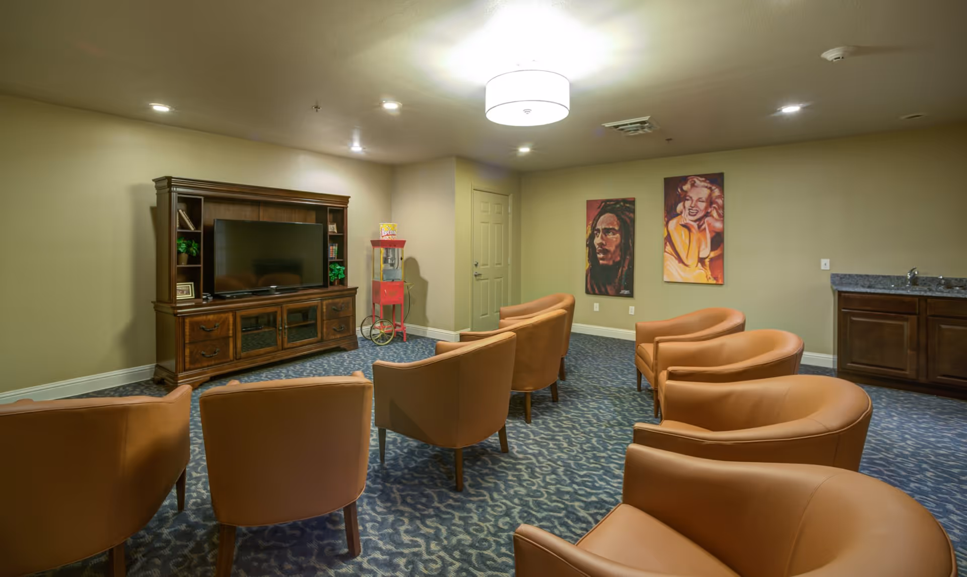A cozy common room with six tan leather chairs arranged in two rows facing a large wooden entertainment center with a flat-screen TV. The room has patterned blue carpet, beige walls, two colorful portrait paintings on one wall, a popcorn machine in the corner, and a small kitchenette with a sink and cabinets.