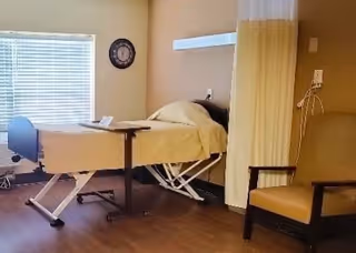 A nursing facility room with a hospital bed covered in beige bedding, a small overbed table, a beige armchair, a wall clock, and a window with blinds. There is a privacy curtain partially drawn and medical equipment mounted on the wall.
