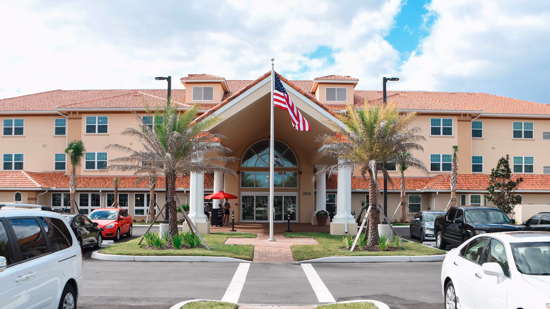 Front exterior view of Shell Harbor Retirement Community building with a large entrance featuring white columns, an American flag on a flagpole in the center, palm trees, and several parked cars in the driveway.