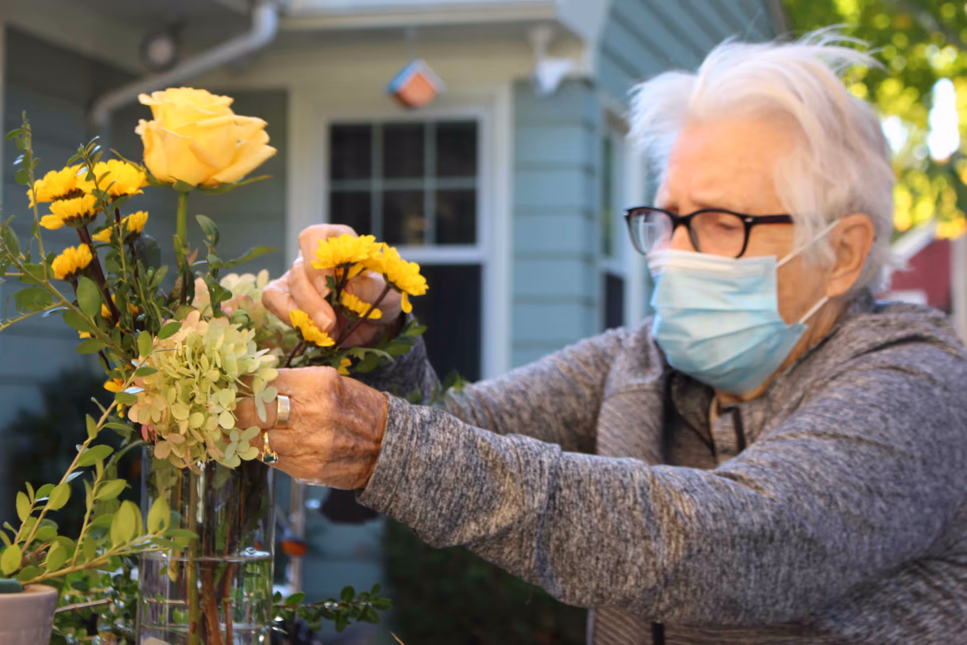 An elderly person wearing glasses and a face mask arranges a bouquet of yellow flowers and greenery in a clear glass vase outdoors, with a house visible in the background.