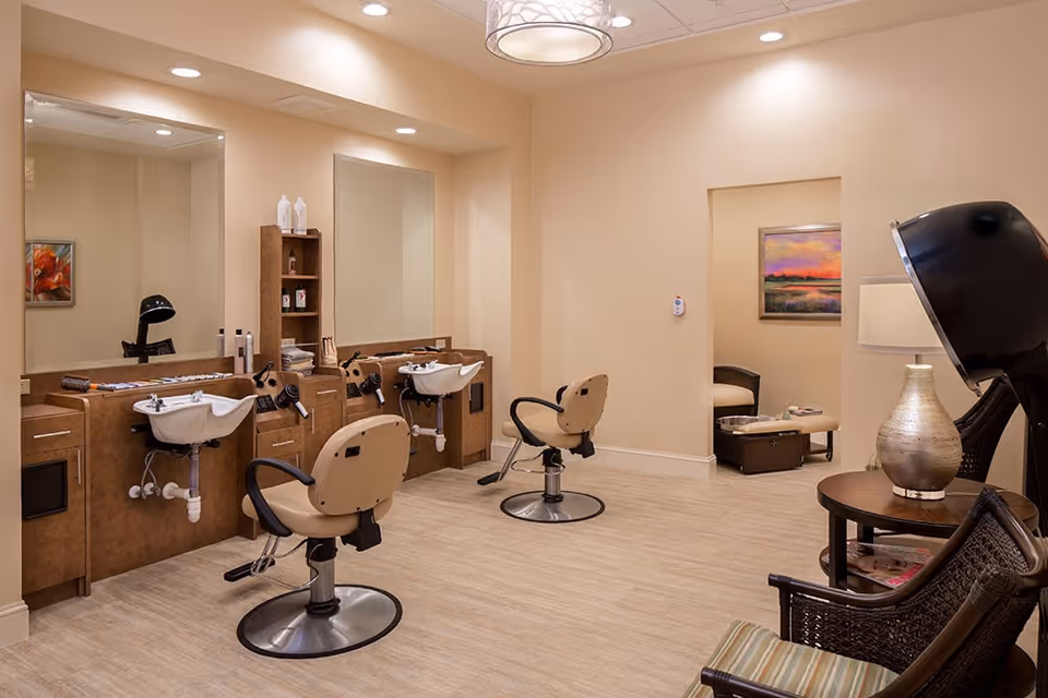 Interior view of a hair salon area in a senior living facility with two salon chairs in front of washbasins and mirrors. The room has beige walls and wood-like flooring. There is a small seating area with a lamp and chairs, and a painting on the wall in an adjoining room.
