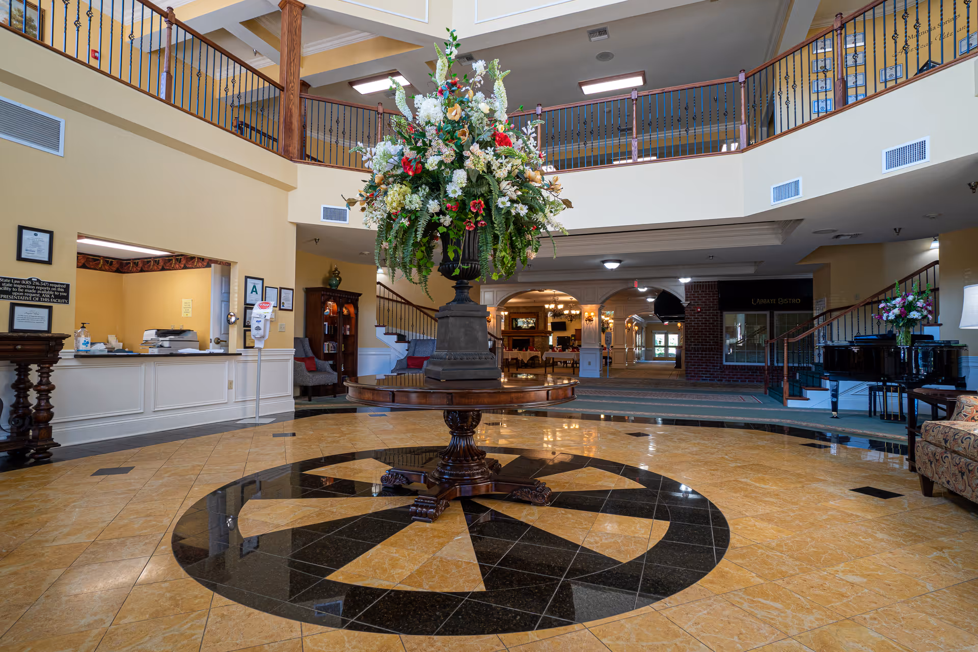 Spacious and elegant lobby area with a large floral arrangement on a round wooden table in the center. The floor features a decorative tile pattern. There is a reception desk to the left, a grand piano to the right, and a staircase leading to an upper balcony. The space is well-lit with natural and artificial lighting, and there are comfortable seating areas visible in the background.