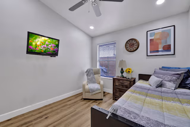 Small furnished bedroom with a bed, nightstand and lamp, an armchair by a window, a wall-mounted TV, ceiling fan, and hardwood floors.