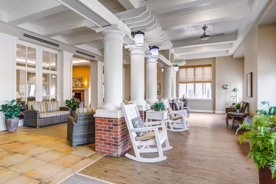 A bright and spacious common area in a senior living facility featuring white rocking chairs with cushions lined up along a wooden floor next to large white columns with brick bases. The room has a high ceiling with exposed beams and ceiling fans. There are wicker armchairs and sofas with striped cushions in an adjacent seating area, potted plants, and large windows with woven shades allowing natural light to fill the space.