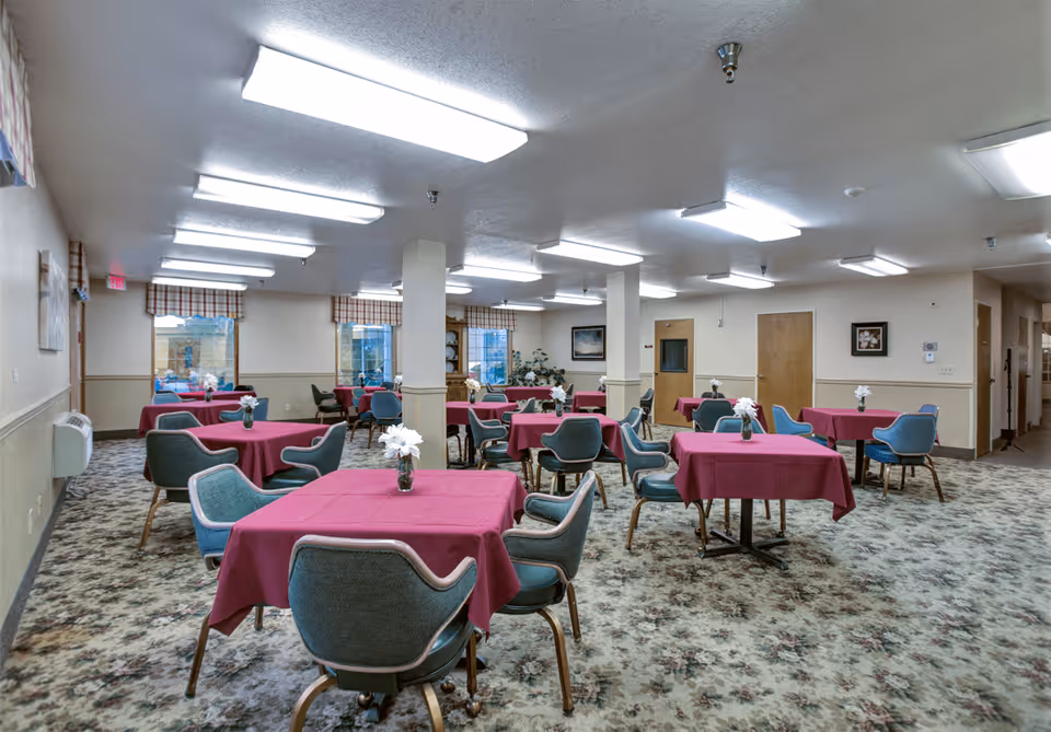 A spacious dining room with multiple tables covered in red tablecloths, each table surrounded by blue cushioned chairs. The room has patterned carpet flooring, beige walls with a chair rail, and several windows with plaid valances. Fluorescent ceiling lights illuminate the space, and small flower arrangements are placed on each table.