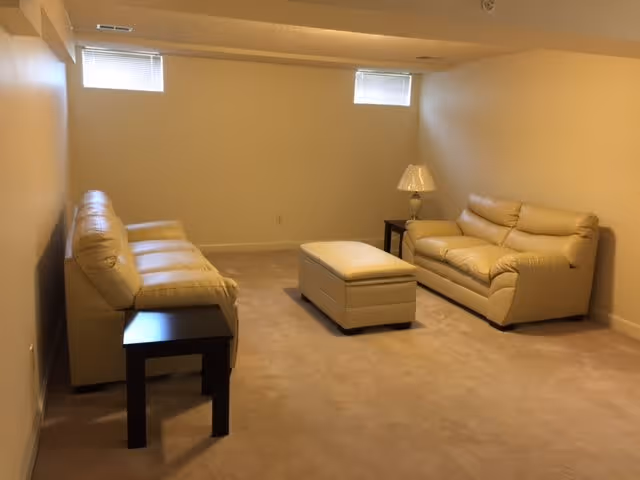A small living room with beige walls and carpet, featuring two beige leather sofas facing each other, a matching ottoman in the center, a dark wooden side table, and a lamp on a small table near one sofa. Two small windows near the ceiling provide natural light.