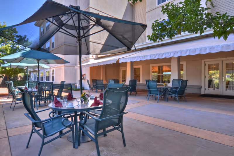 Outdoor patio area at a senior living facility with multiple round tables and chairs. Each table has a large umbrella for shade and is set with cups, glasses, and folded napkins. The patio is adjacent to a building with windows and doors, and there are some trees and greenery visible in the background.