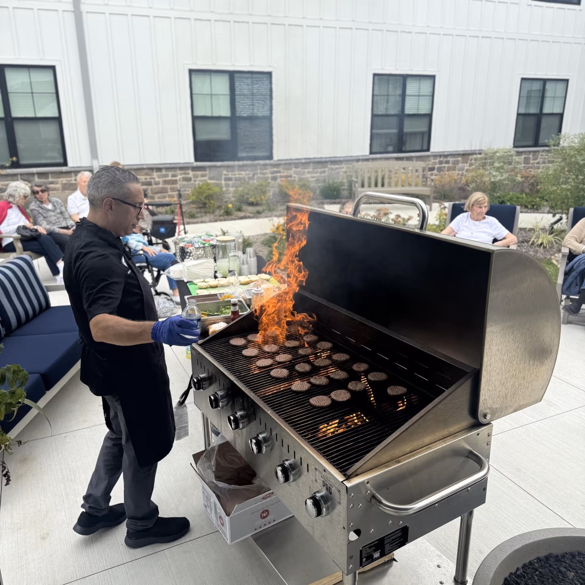 A man wearing gloves and an apron is grilling multiple burger patties on a large outdoor grill with flames visible. In the background, several elderly people are seated on outdoor furniture in a courtyard area with a building wall and windows behind them.