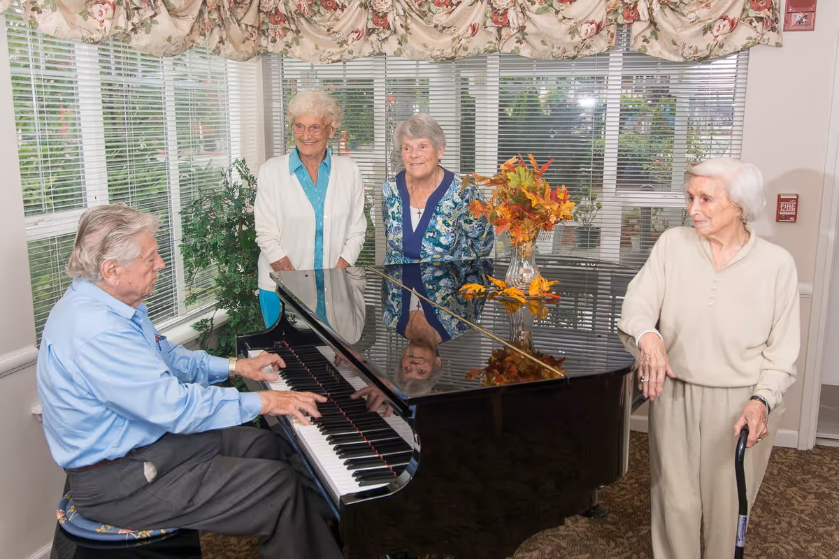 Four elderly residents gathered around a grand piano in a bright sitting room, with one man playing and three women standing nearby.