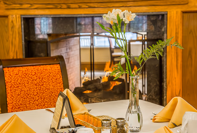 A dining table set with folded yellow napkins, salt and pepper shakers, a wicker basket, and a glass vase holding white flowers and green leaves. In the background, there is a lit fireplace with a wooden mantel and a chair with an orange patterned cushion.