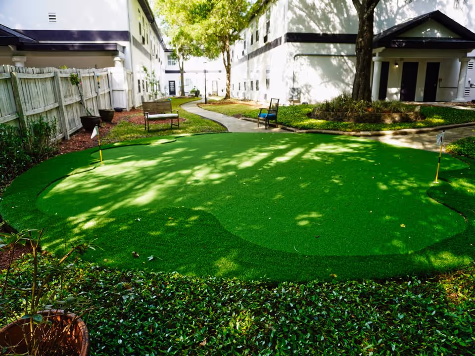 Outdoor putting green area with two small flags, surrounded by greenery and benches. The area is located between two white buildings with windows, and a paved walkway runs through the space. Trees provide shade over the putting green.