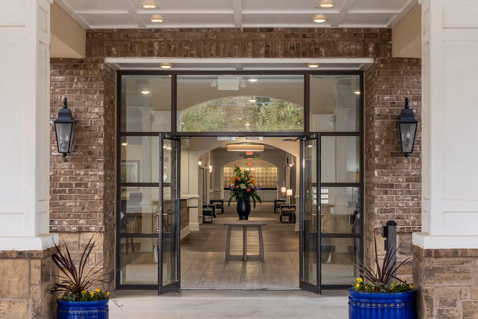 Entrance to a building with double glass doors open, flanked by brick walls and black lantern-style lights. Two blue planters with plants and flowers are positioned on either side of the entrance. Inside, a hallway with a table holding a large floral arrangement is visible, leading to a well-lit interior space.
