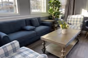 A cozy living room area with a dark blue sofa, two plaid armchairs, a wooden coffee table with a small potted plant, large windows letting in natural light, and a tall green plant in the corner.