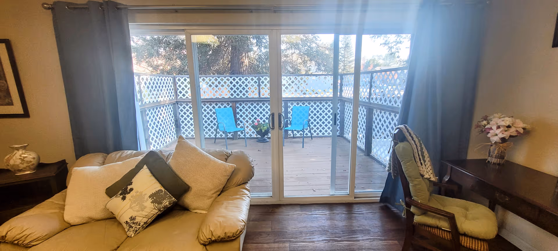 Interior view of a senior living facility room with a beige cushioned armchair with pillows on the left, a wooden desk with a green cushioned chair and a vase of flowers on the right, and large sliding glass doors leading to a small outdoor deck with two blue chairs and a potted plant. The deck is enclosed with white lattice fencing.