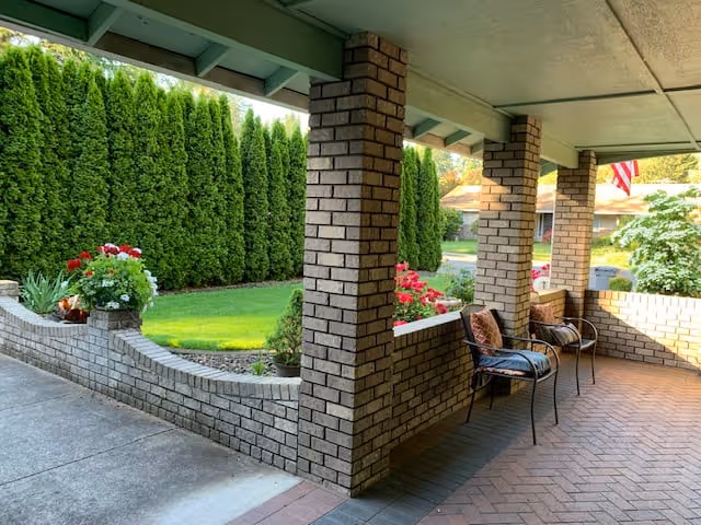 Covered outdoor patio area with brick pillars and low brick walls, featuring two metal chairs with cushions. The patio overlooks a well-maintained green lawn bordered by tall evergreen trees and colorful flower beds.