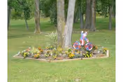 A small garden bed with various plants and flowers surrounded by grass, featuring a decorative pink and blue tricycle placed upright near the center, with trees in the background.