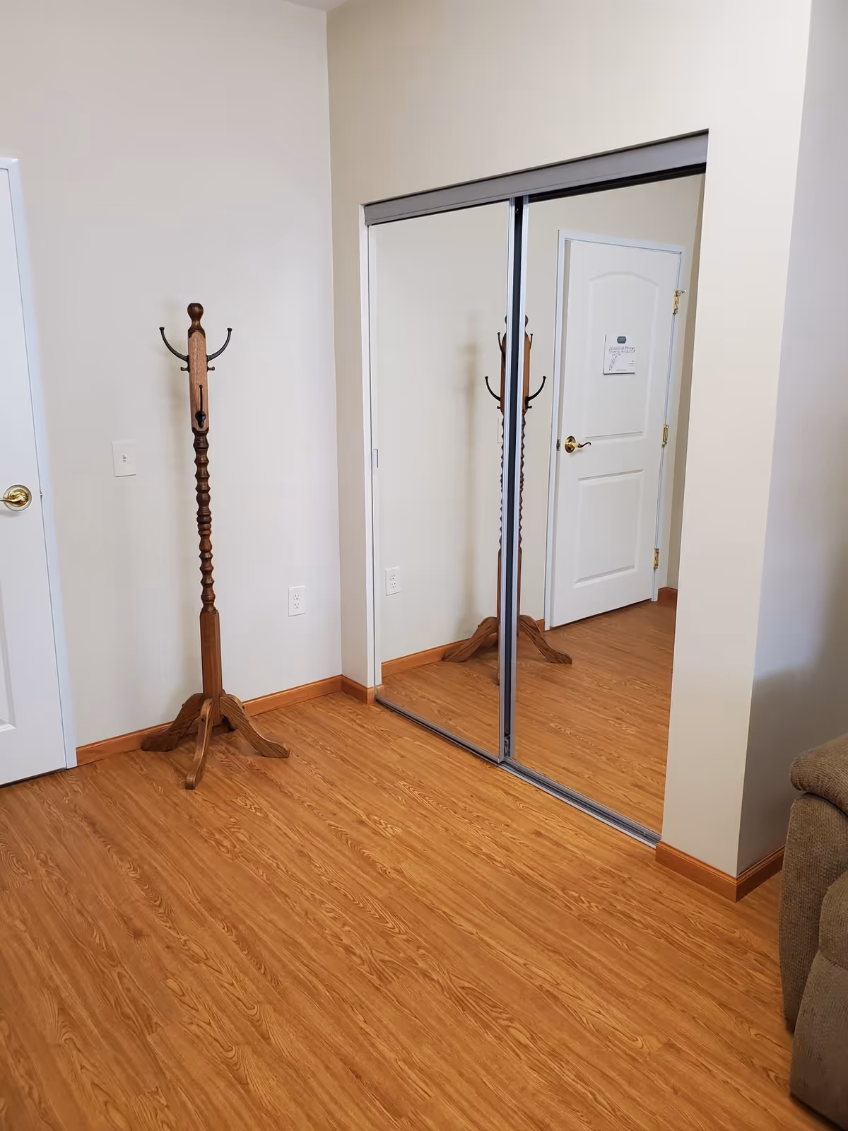 Interior corner of a room with wood flooring, a wooden coat rack, a white door with a gold handle, and a closet with sliding mirrored doors reflecting the coat rack and door.