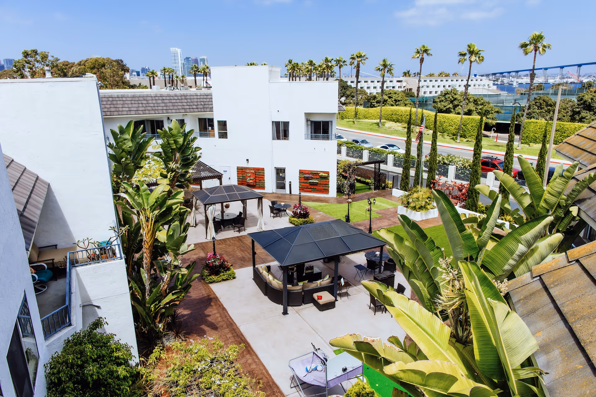 Aerial view of an outdoor courtyard area at Coronado Retirement Village featuring white buildings, palm trees, two covered seating areas with chairs and tables, landscaped greenery, and a distant view of a bridge and water.