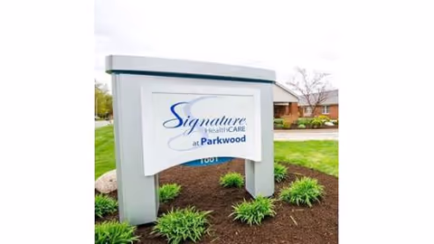 Outdoor view of a white and gray sign for Signature HealthCARE at Parkwood, surrounded by green plants and mulch, with a building and trees in the background.
