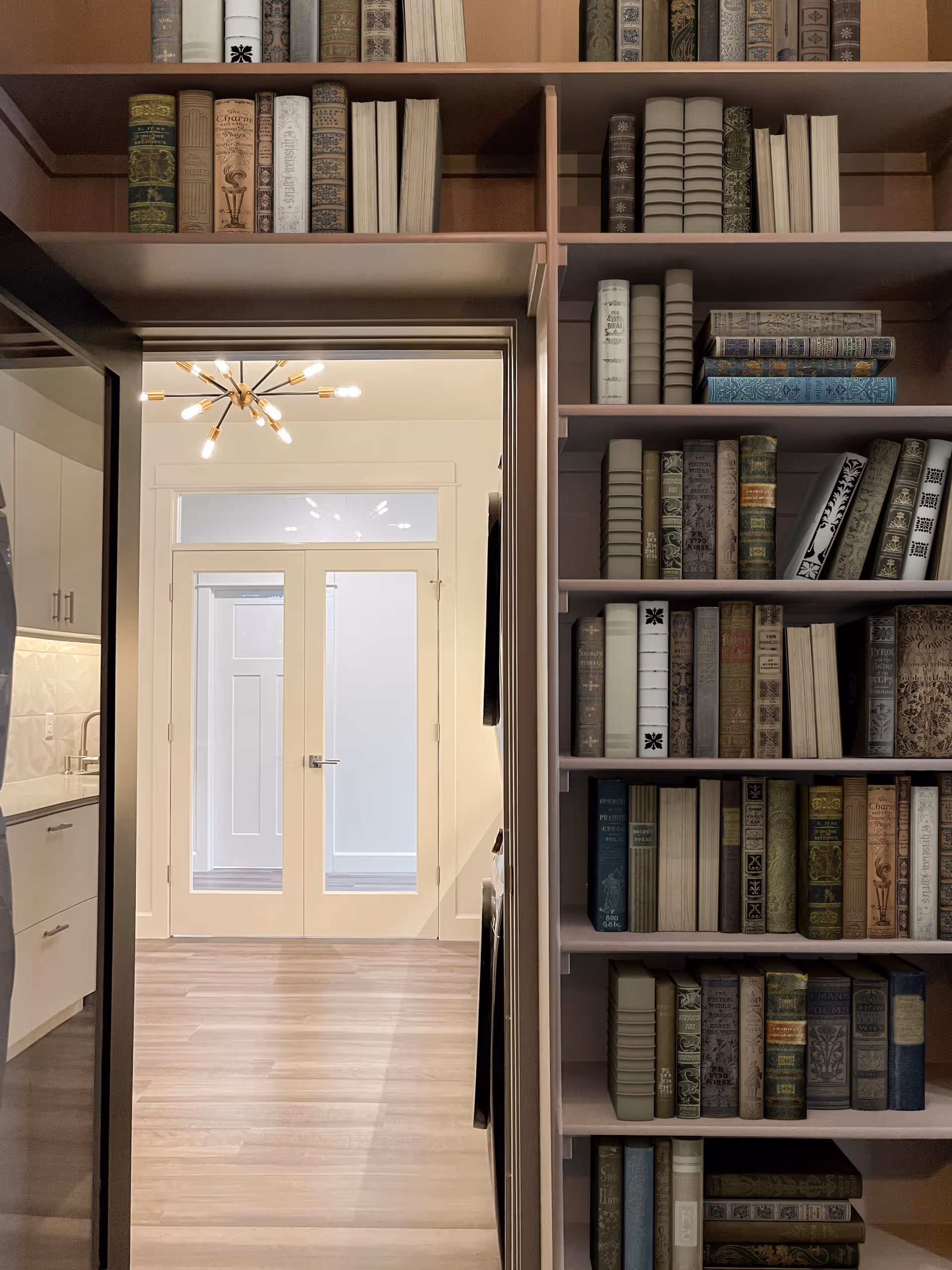 Interior view showing a built-in bookshelf filled with various books on the right side, a doorway leading to a bright room with double glass doors and a modern chandelier, and part of a kitchen area with white cabinets and a countertop on the left.