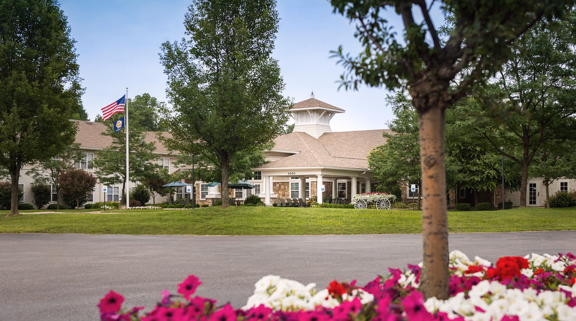 Exterior view of The Addison of Park Crescent building with a driveway in front, green lawn, trees, and colorful flowers in the foreground. Two flags are flying on a flagpole near the entrance.