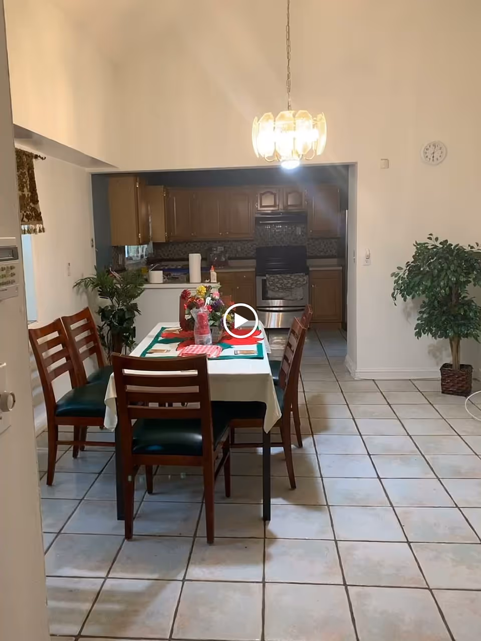 A dining area with a rectangular table covered with a white tablecloth and a green runner, set with placemats and a centerpiece of flowers. Six wooden chairs with dark green cushions surround the table. The dining area is adjacent to a kitchen with wooden cabinets, a stove, and a tiled backsplash. The floor is tiled with large square tiles, and there are two potted plants in the room. A hanging light fixture is above the table, and a clock is mounted on the wall.