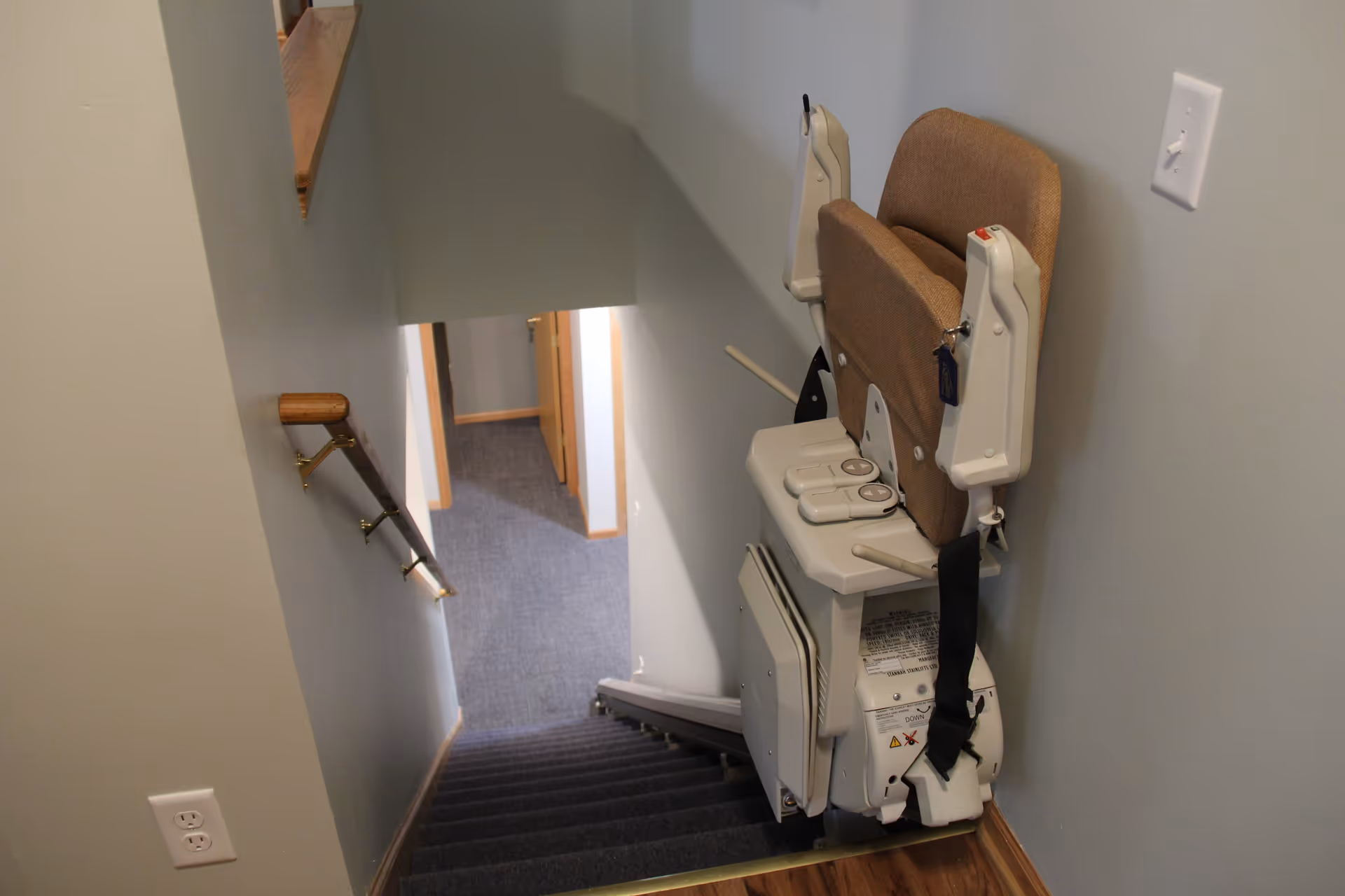 A stairway with a beige stair lift chair installed on the right side. The stair lift has control buttons on the armrest and a safety belt. The stairs are carpeted in dark gray, and there is a wooden handrail on the left wall. The walls are painted light gray, and there is a white electrical outlet near the bottom of the stairs.