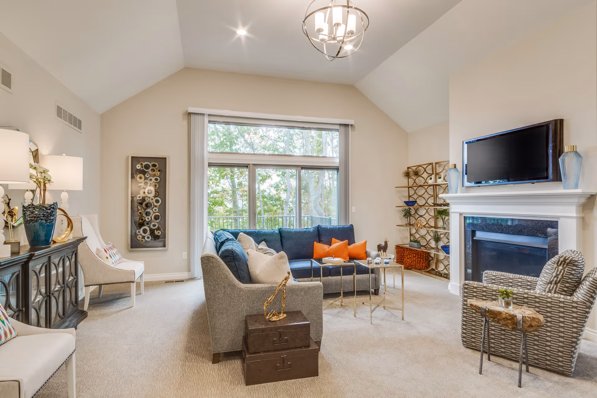 A bright and spacious living room with a vaulted ceiling, large window showing trees outside, a gray sectional sofa with orange and beige pillows, a patterned armchair, a fireplace with a mounted TV above it, decorative shelves, and a console table with lamps and decor.