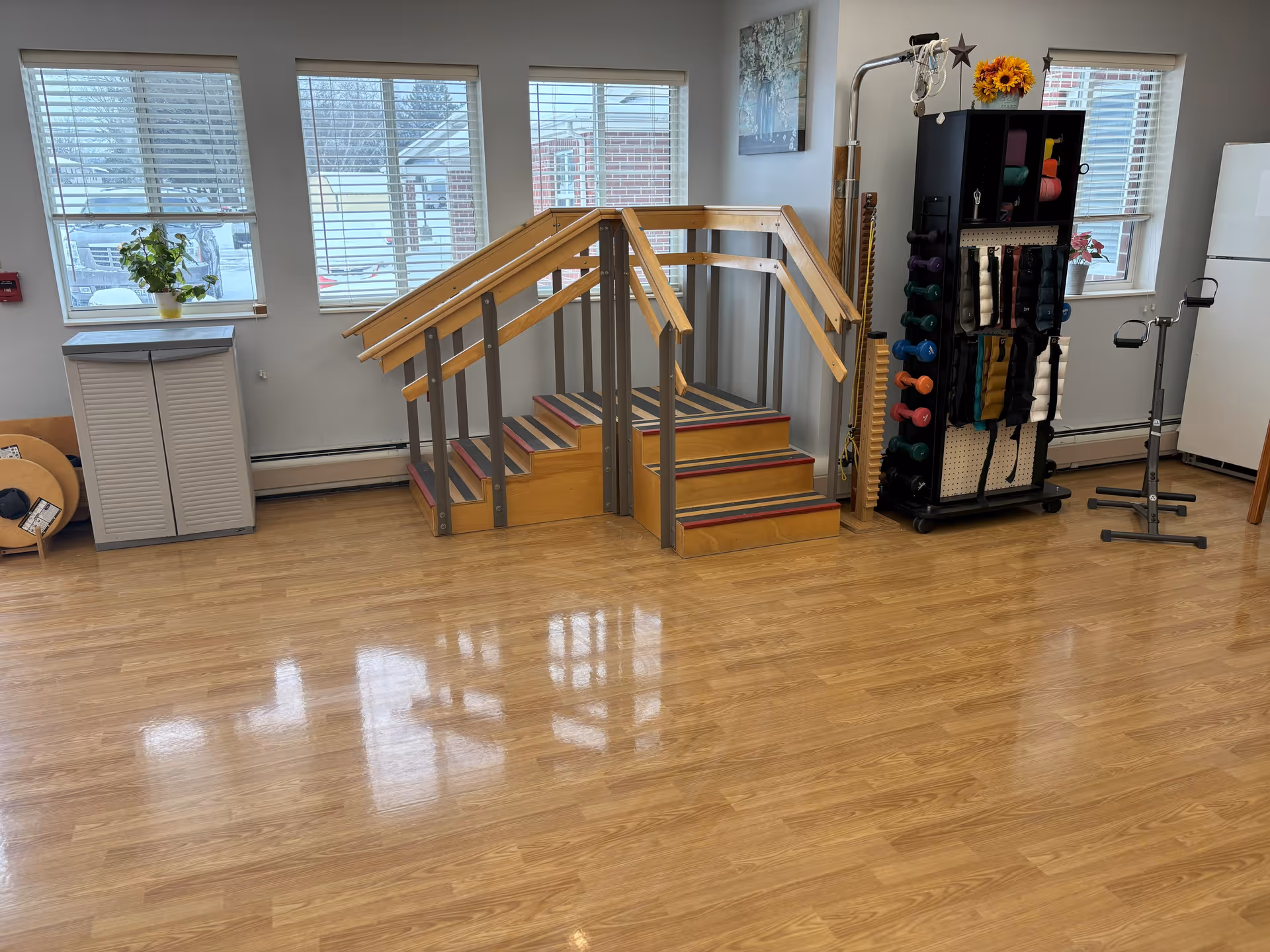 A rehabilitation or physical therapy room with wooden handrails and steps for exercise, a rack holding various exercise weights and bands, a white refrigerator, and windows with blinds letting in natural light.