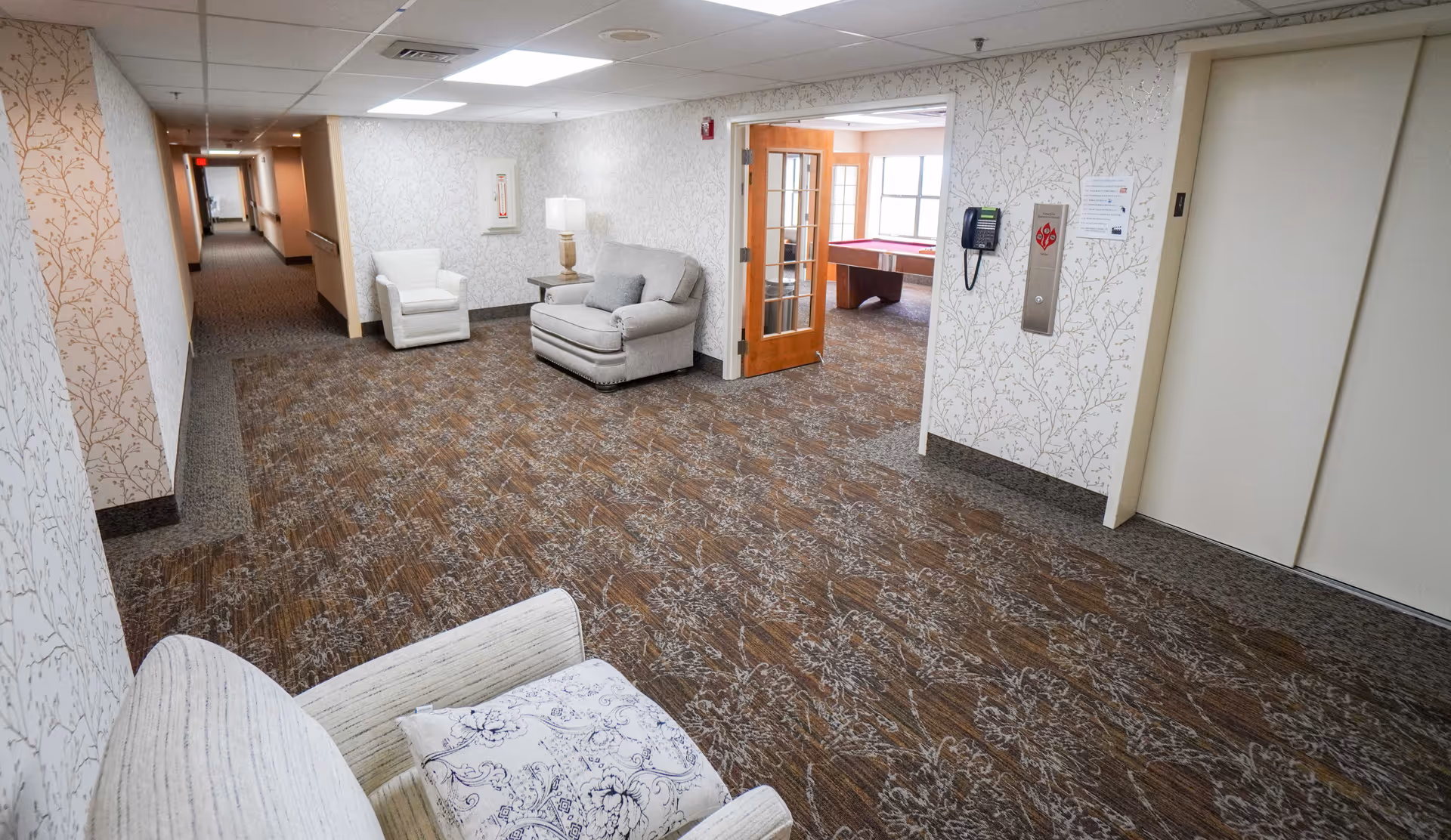 Carpeted hallway/common area in a senior living facility with seating, elevator doors, and a doorway leading to a game room with a pool table.