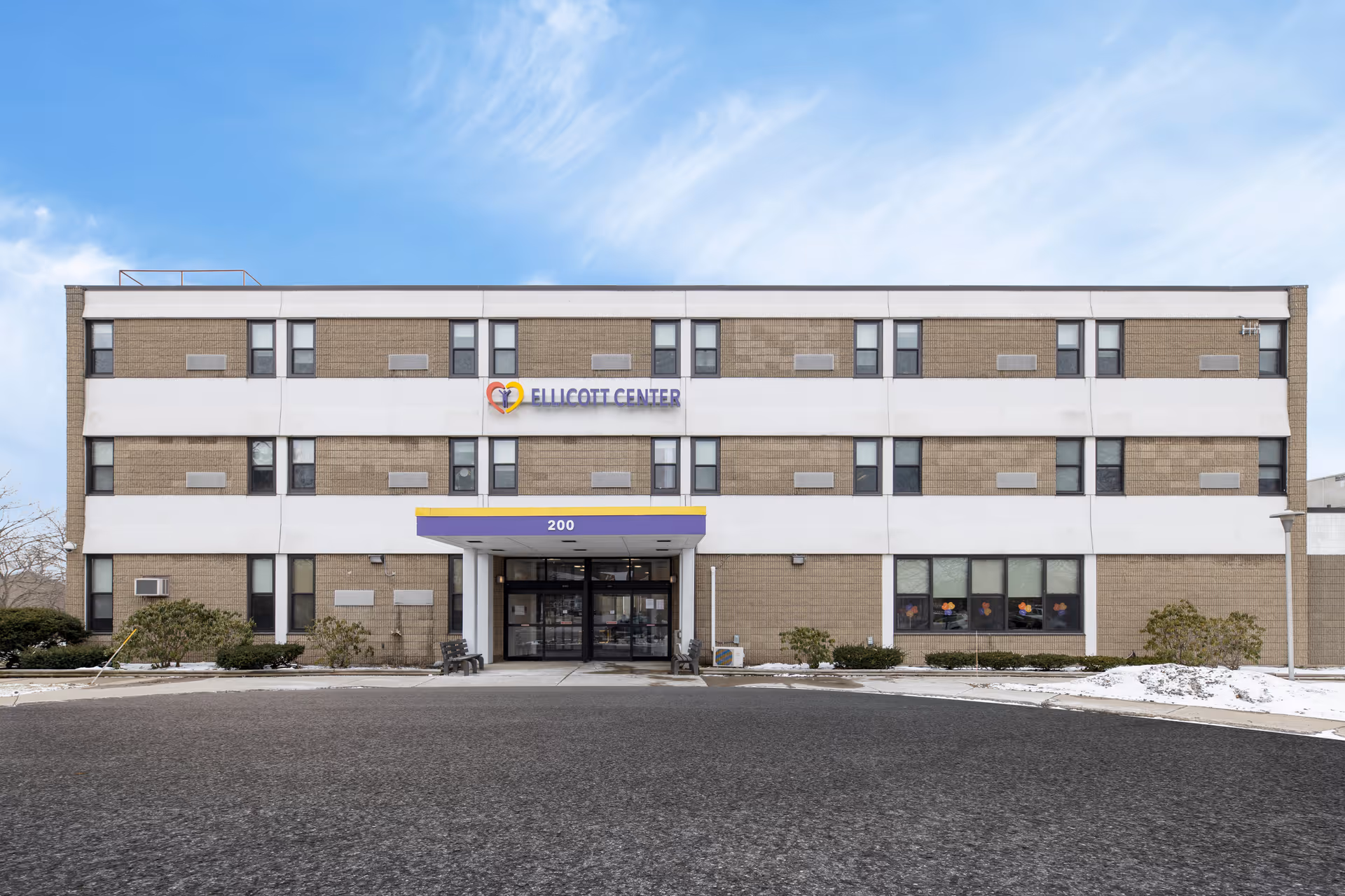 Front exterior view of the Ellicott Center building, a three-story structure with a beige and white facade, multiple windows, a main entrance with a purple and yellow awning displaying the number 200, and some bushes and snow on the ground.