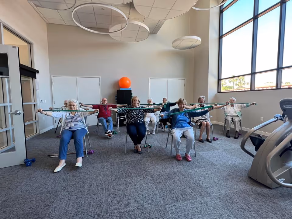 A group of elderly individuals seated in chairs in a spacious, well-lit exercise room performing a stretching exercise with resistance bands. The room has large windows, exercise equipment, and modern ceiling light fixtures.