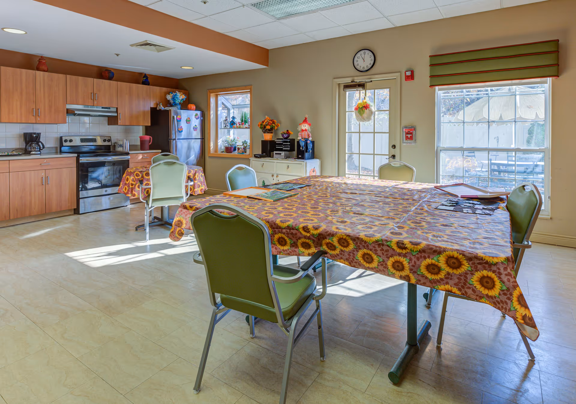 A bright kitchen and dining area with wooden cabinets, a stainless steel stove and refrigerator, and tables covered with sunflower-patterned tablecloths. Green chairs surround the tables, and there are windows and a glass door letting in natural light. Decorative items and plants are placed on a white cabinet and windowsill.