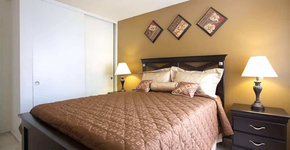 A bedroom with a large bed covered in a brown quilted bedspread and multiple pillows. There are two bedside tables, each with a lamp. The wall behind the bed is painted a warm beige color and decorated with three square floral art pieces arranged diagonally. A white sliding closet door is visible on the left side of the room.