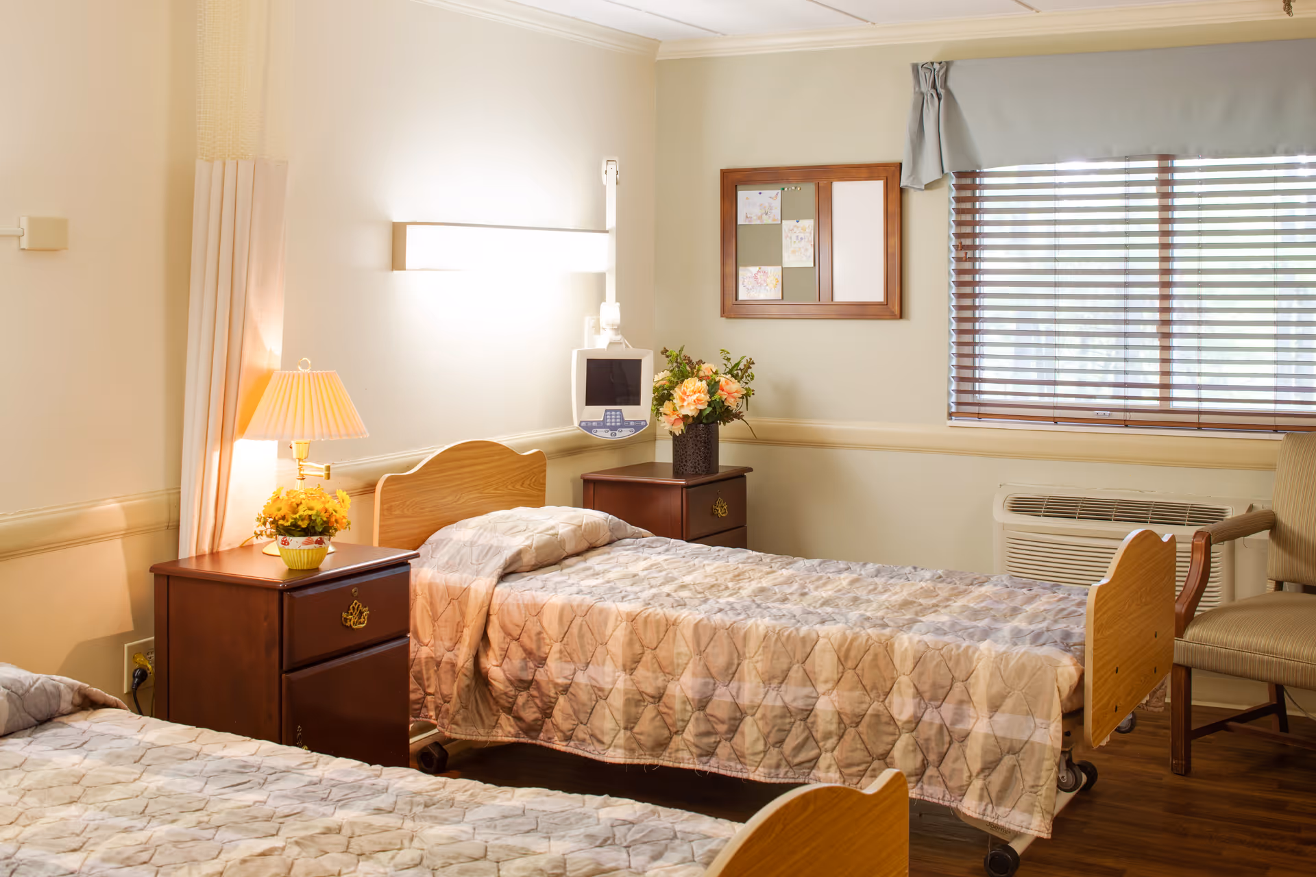 A well-lit senior living bedroom with two single beds covered in patterned quilts. Between the beds is a wooden nightstand with a lamp and a small flower pot. On the wall above the nightstand is a mounted medical monitor. A window with wooden blinds and a chair are visible on the right side. The room has light-colored walls and wooden flooring.