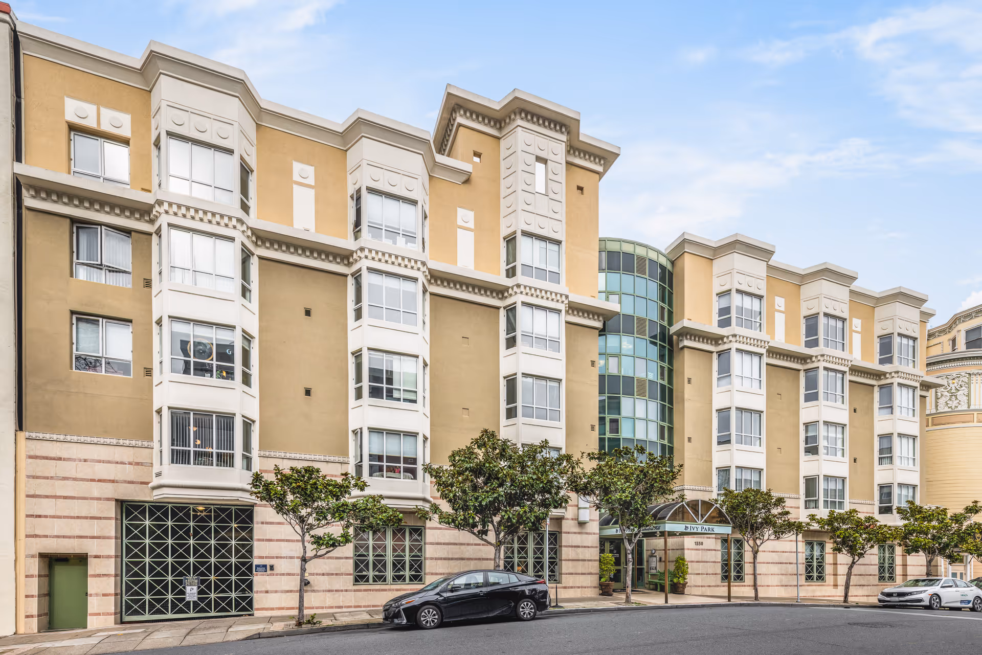 Exterior view of a multi-story senior living facility named Ivy Park at Cathedral Hill, featuring beige and white facade with large windows, a glass entrance, and several small trees along the sidewalk. Two cars are parked on the street in front of the building under a partly cloudy sky.