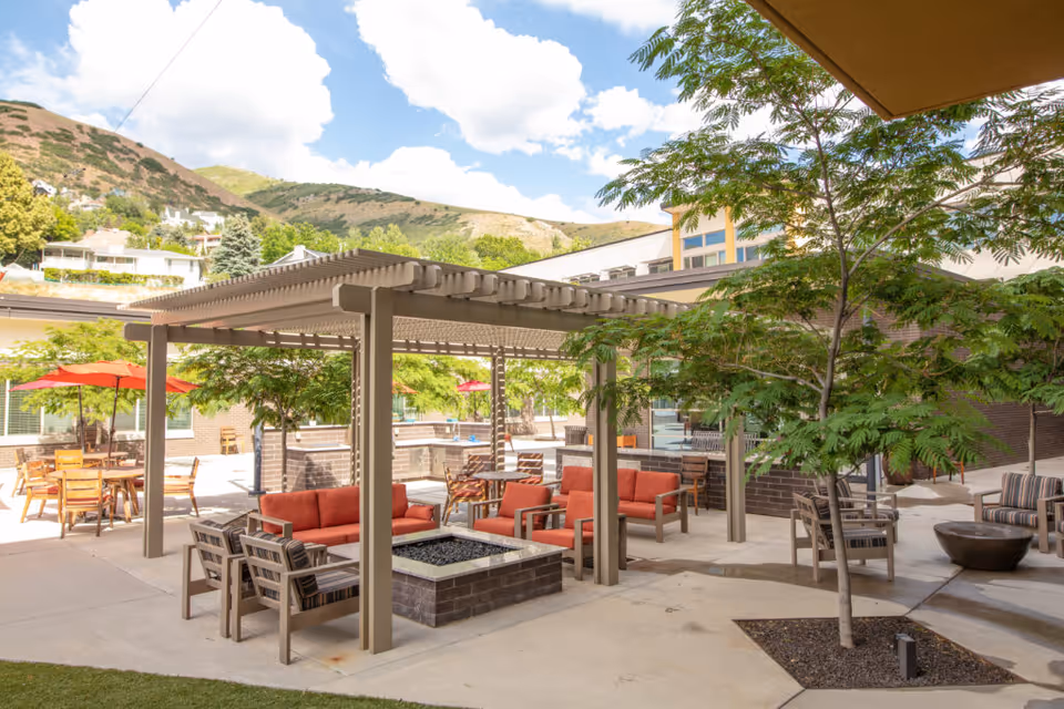 Outdoor seating area at The Ridge Foothill featuring cushioned chairs and sofas under a pergola, surrounded by trees and tables with red umbrellas, with hills and a partly cloudy sky in the background.