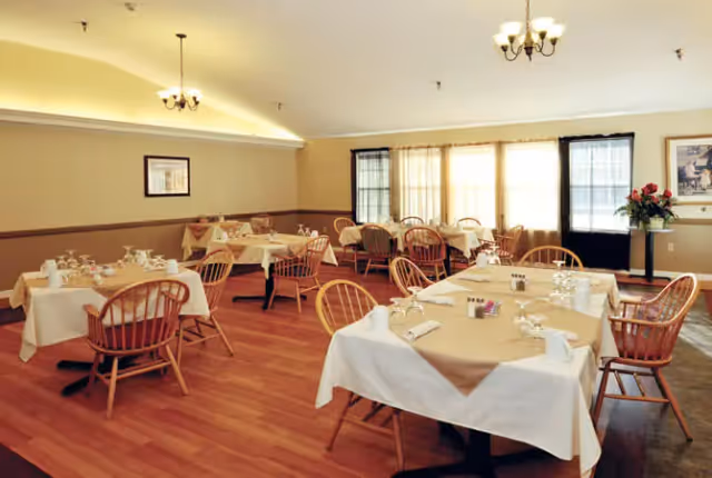 A dining room with several tables covered in white and beige tablecloths, each set with glassware, napkins, and condiments. Wooden chairs surround the tables. The room has wooden flooring, beige walls with a chair rail, and large windows with sheer curtains letting in natural light. There are two framed pictures on the walls and a small table with a flower arrangement near the windows.