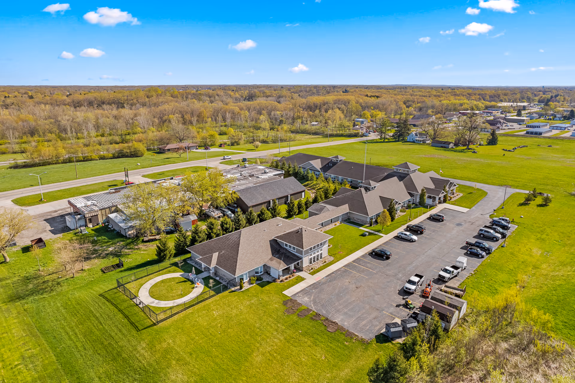 Aerial view of Woodland Terrace of Paw Paw Lake facility showing multiple connected buildings with pitched roofs, a parking lot with several cars, a fenced circular outdoor area, and surrounding green lawns and trees under a blue sky with scattered clouds.